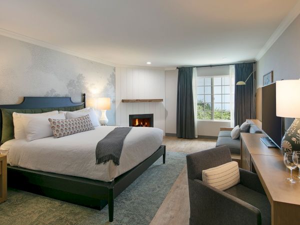 A spacious hotel room with a large bed, two nightstands, a sitting area, fireplace, and a work desk near a window with blue curtains.